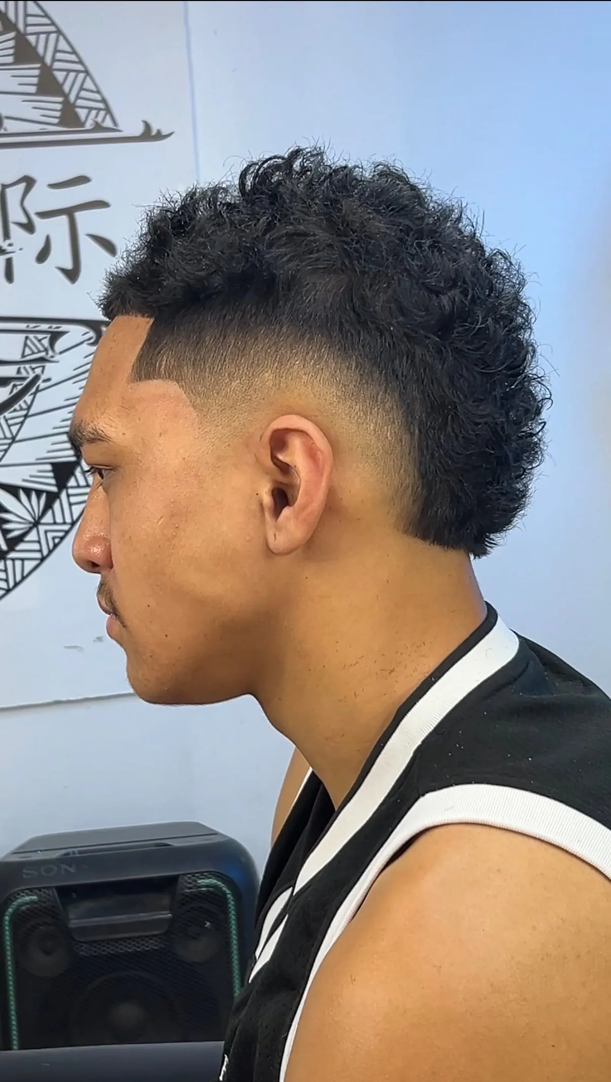Side profile of a curly mullet over a high skin fade, with a shaved part line cut in at the temple.